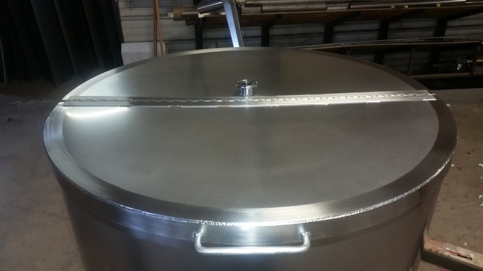 Large-diameter stainless steel tank with a hinged lid, sanitary ferrule and lifting handle on the shop floor